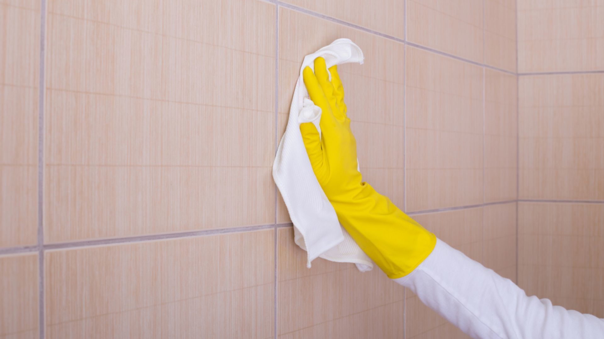 バスルームで、黄色い掃除用手袋をはめた手でタイル張りの壁を白い布で拭いています。