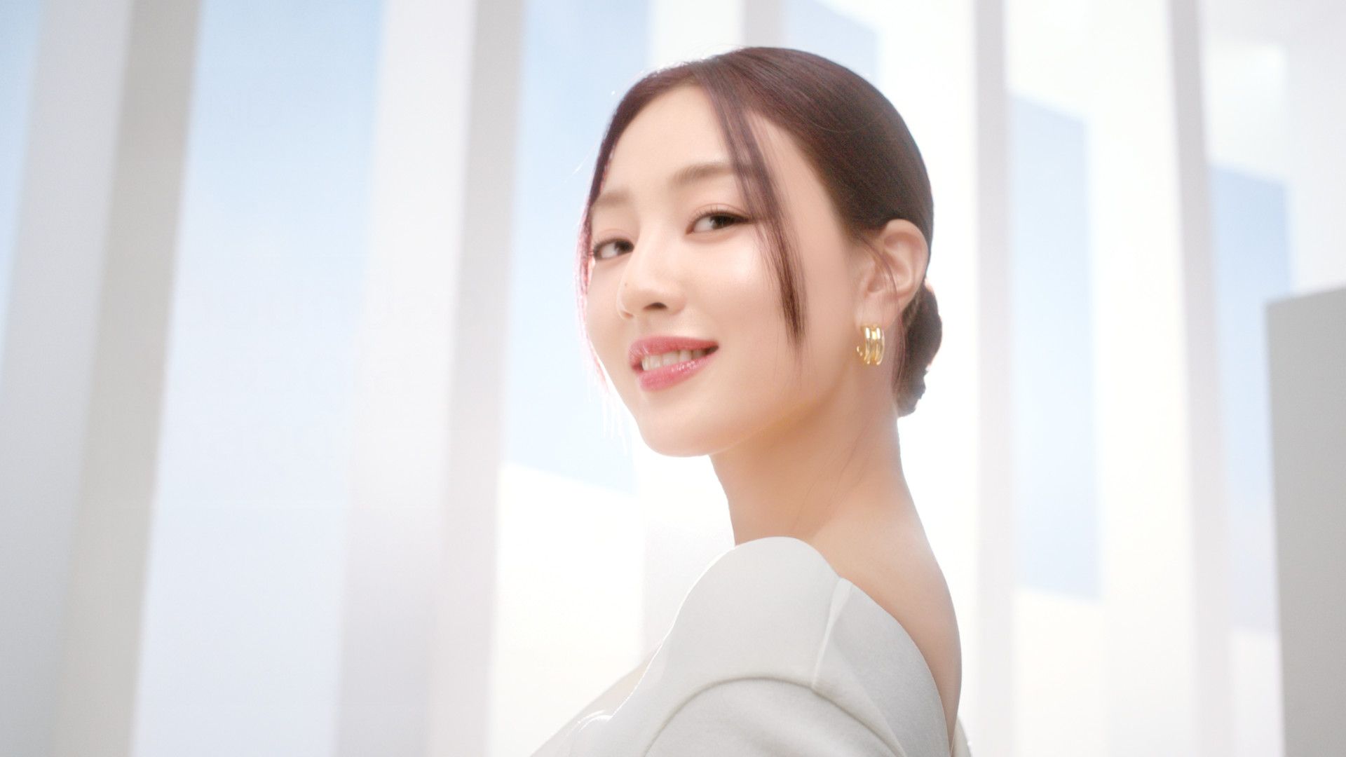 なめらかなヘアスタイルとエレガントなイヤリングをつけた人物が、明るく柔らかな光とモダンな建築に囲まれています。